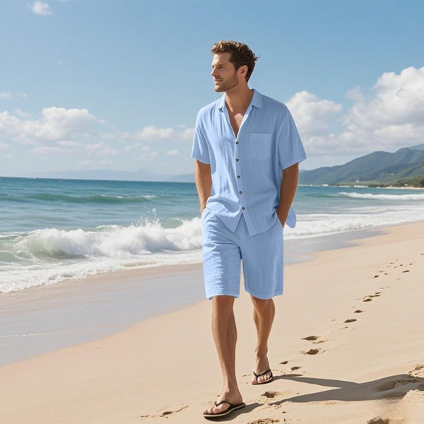 Man in light blue outfit standing on a beach with ocean and mountains in the background